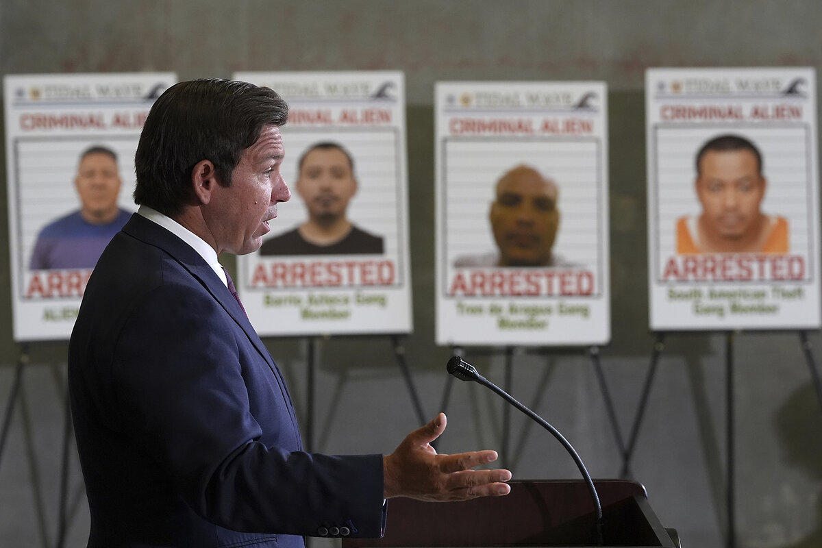 Gov. Ron DeSantis speaks to reporters. Next to him are large posters of suspected criminals caught in recent immigration strikes..